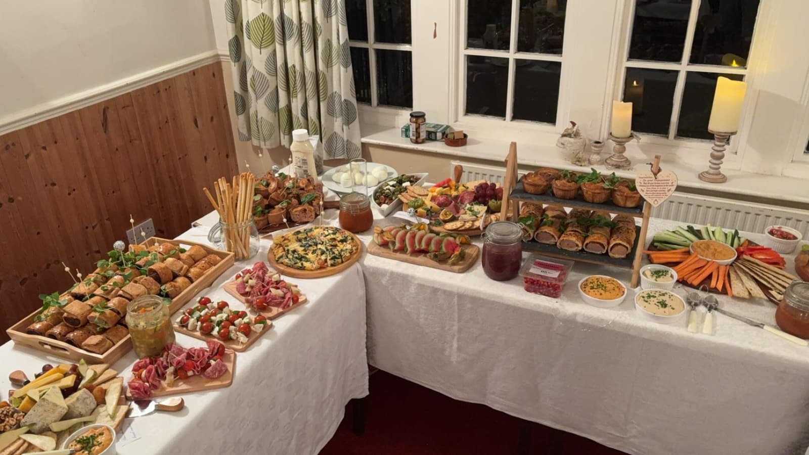 A buffet across two tables with sausage rolls, pork pies on a tiered stand, a frittata, skewers, crudités and dips