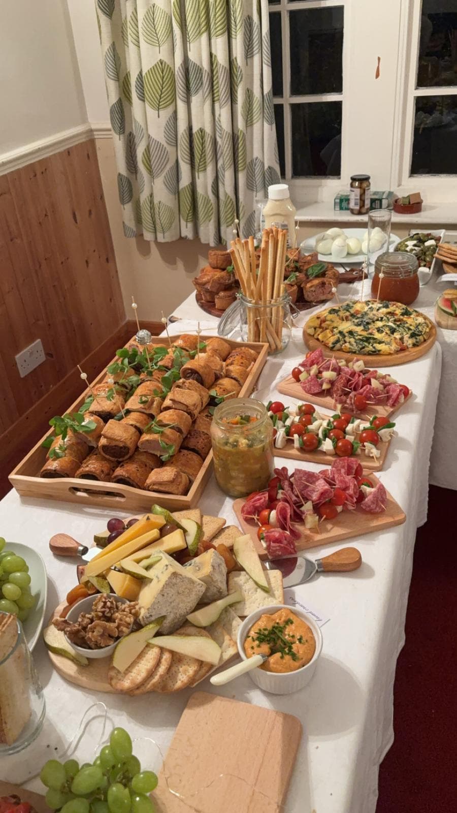 A buffet table laid with sausage rolls, a cheese board, cured meat skewers and a spinach frittata